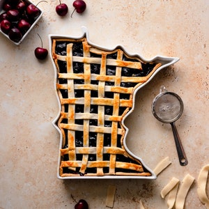 Könnte beinhalten: Ein Kirschkuchen, gebacken in einer weißen Keramikform in Form des Bundesstaates Minnesota. Der Kuchen hat eine Gitterkruste und ist von frischen Kirschen umgeben.