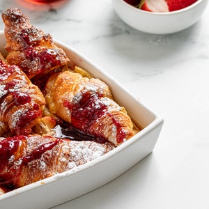 May include: A white ceramic baking dish filled with baked French toast topped with raspberry sauce and powdered sugar. The dish is on a white marble surface.