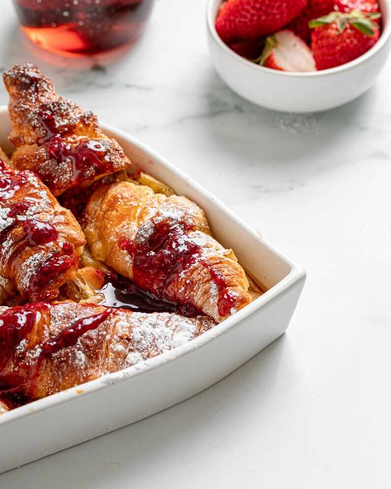 May include: A white ceramic baking dish filled with baked French toast topped with raspberry sauce and powdered sugar. The dish is on a white marble surface.