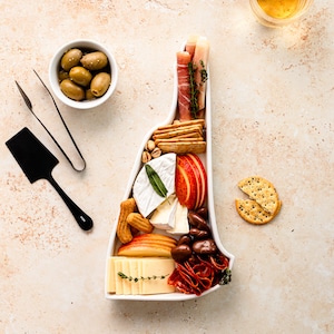 May include: A white ceramic serving tray shaped like the state of Virginia, filled with a variety of snacks, including cheese, crackers, fruit, salami, and olives.