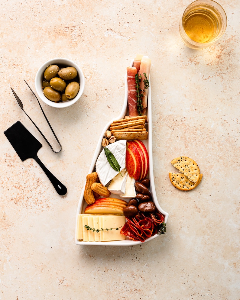 May include: A white ceramic serving tray shaped like the state of Virginia, filled with a variety of snacks, including cheese, crackers, fruit, salami, and olives.