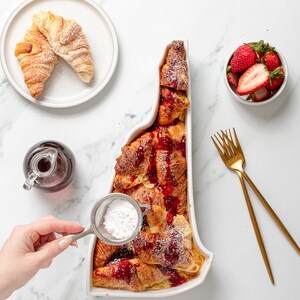 May include: A white ceramic baking dish shaped like the state of New Hampshire filled with a baked French toast casserole topped with a red berry sauce and powdered sugar. A hand is sprinkling powdered sugar over the casserole.