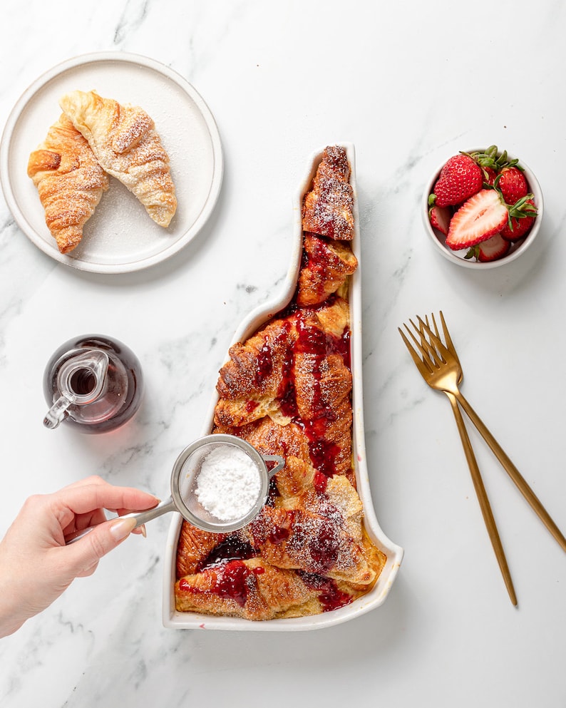 May include: A white ceramic baking dish shaped like the state of New Hampshire filled with a baked French toast casserole topped with a red berry sauce and powdered sugar. A hand is sprinkling powdered sugar over the casserole.