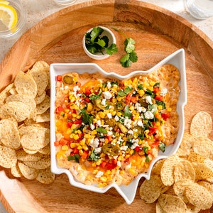 Könnte beinhalten: Eine weiße, Ohio-förmige Servierschale gefüllt mit einem cremigen Dip, belegt mit Mais, roten Paprika, Jalapeños und Käse. Tortilla-Chips umgeben die Schale auf einem Holztablett, mit Koriander und Zitronenwasser in der Nähe. Eine festliche Vorspeise.