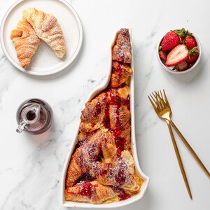 May include: A white ceramic baking dish shaped like the state of Vermont filled with a sweet bread pudding made with croissants, topped with raspberry sauce and powdered sugar. A small white bowl filled with fresh strawberries sits on a white marble surface.