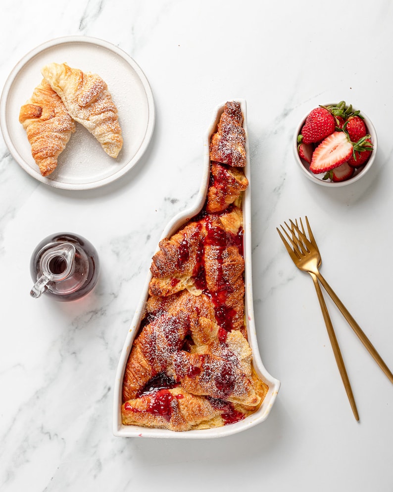 May include: A white ceramic baking dish shaped like the state of Vermont filled with a sweet bread pudding made with croissants, topped with raspberry sauce and powdered sugar. A small white bowl filled with fresh strawberries sits on a white marble surface.