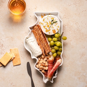Könnte beinhalten: Eine weiße Keramik-Servierplatte in Form des Bundesstaates Illinois, gefüllt mit einem Käseteller mit verschiedenen Käsesorten, Crackern, Trauben, Mandeln und Prosciutto.