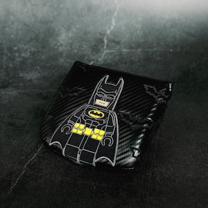 Puede incluir: Una cartera negra de dos pliegues con textura de fibra de carbono y un gráfico bordado de una figura estilizada de Batman. El diseño incluye un emblema amarillo en el pecho y un cinturón utilitario amarillo, con sutiles siluetas de murciélagos.