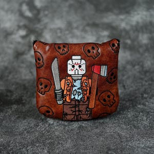 Könnte beinhalten: Braune Golfschlägerhaube aus Leder mit einem gestickten Design eines Charakters mit Hockeymaske, der eine Machete und eine Axt hält. Die Haube ist mit Totenkopfmotiven verziert.