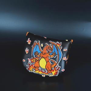 May include: Black putter cover featuring a vibrant embroidered image of a Charizard character surrounded by flames. The cover has orange trim and a magnetic closure. A unique accessory for golf enthusiasts.