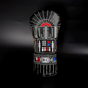 Puede incluir: Funda negra para la cabeza del palo de golf con un diseño inspirado en un popular personaje de ciencia ficción. La funda presenta un diseño bordado detallado con detalles en rojo, blanco, gris y azul. La parte superior tiene un diseño de sol.