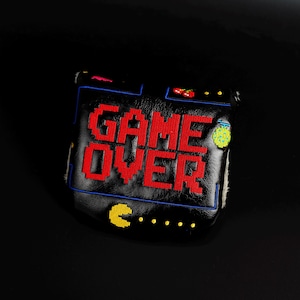 May include: Black golf club head cover with the words "GAME OVER" in red pixelated letters. The cover features a retro video game design with Pac-Man and other game elements. The cover is made of a leather-like material.