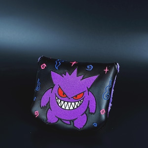May include: Black golf club head cover featuring a purple embroidered Gengar character with red eyes and a wide grin. The cover has a black background with embroidered swirls and plus signs. The edges are lined with purple fur.