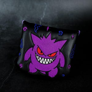 May include: A black putter headcover featuring an embroidered purple creature with red eyes and a wide, toothy grin. The design includes small purple and blue swirling accents and star shapes scattered across the surface of the black material.