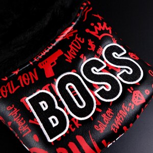May include: Black golf club head cover with the word "BOSS" in large, white-outlined letters. The cover has a red graffiti-style design with crowns, guns, and dollar signs. The interior is lined with black plush material.
