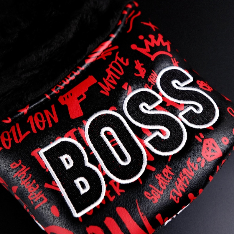 May include: Black golf club head cover with the word "BOSS" in large, white-outlined letters. The cover has a red graffiti-style design with crowns, guns, and dollar signs. The interior is lined with black plush material.