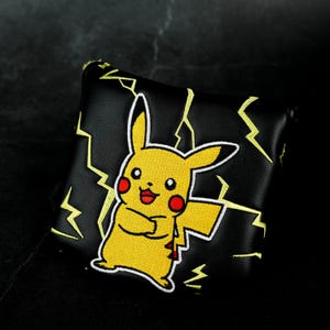Puede incluir: Una funda para putter de golf de color negro y forma cuadrada que presenta un personaje Pikachu amarillo bordado con mejillas rojas y una amplia sonrisa. La funda está decorada con patrones de rayos amarillos cosidos sobre la superficie negra.