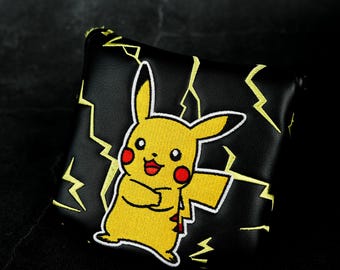 May include: A black square-shaped golf putter headcover featuring an embroidered yellow Pikachu character with red cheeks and a wide smile. The cover is decorated with yellow lightning bolt patterns stitched across the black surface.