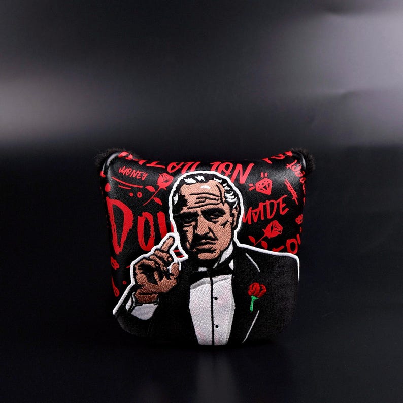 May include: Black golf putter cover featuring a cartoon image of a man in a suit with a red rose. The cover has the word "DON" in red, with other red text and diamond graphics on a black background.