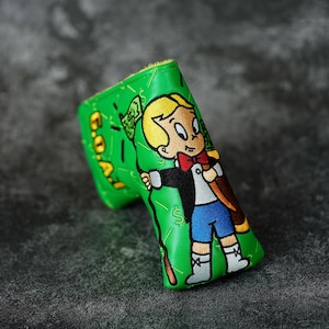May include: A green golf club head cover featuring an embroidered cartoon character holding a money bag. The cover has the text "G.O.A.T." and dollar signs. The character is wearing a black suit, blue shorts, and a red bow tie.