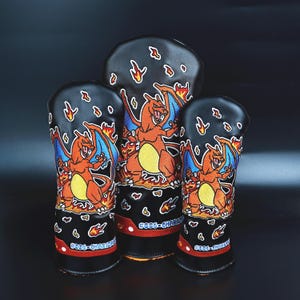 May include: Three black golf club head covers featuring a cartoon Charizard design. The covers have a red and orange trim, with flames and the text "#006-CHARIZARD" printed on them. The Charizard is orange with blue wings.