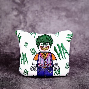 May include: White golf ball marker with an embroidered image of the Joker from the Batman franchise. The Joker is wearing a green and purple suit and has a green mohawk. The background is white with the word "HA" repeated in green.