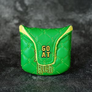 May include: A green golf club head cover with a quilted pattern and yellow trim. The cover features the text "GO AT RICH" in yellow and white, along with dollar signs. The cover is designed to protect golf clubs.