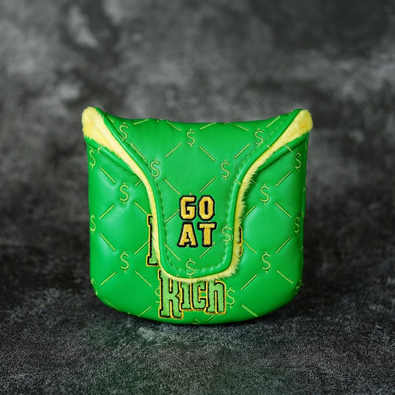 May include: A green golf club head cover with a quilted pattern and yellow trim. The cover features the text "GO AT RICH" in yellow and white, along with dollar signs. The cover is designed to protect golf clubs.