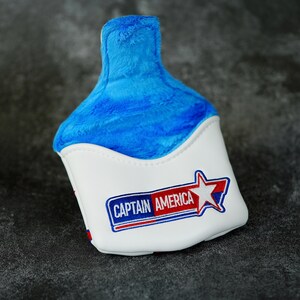 May include: A white and blue golf club head cover featuring the text "CAPTAIN AMERICA" in a red and blue design. The top of the cover is made of blue plush material.