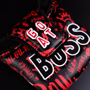 May include: Black golf club head cover with red graffiti-style lettering and the words "GOAT" and "BOSS" in white. The cover has a soft, black interior and a textured exterior.