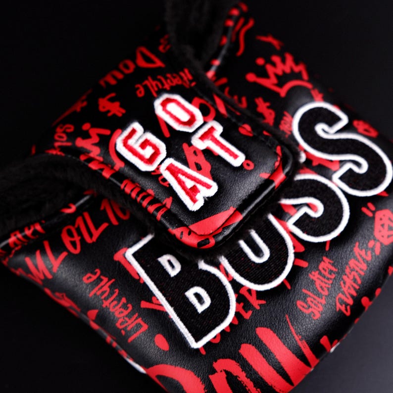 May include: Black golf club head cover with red graffiti-style lettering and the words "GOAT" and "BOSS" in white. The cover has a soft, black interior and a textured exterior.