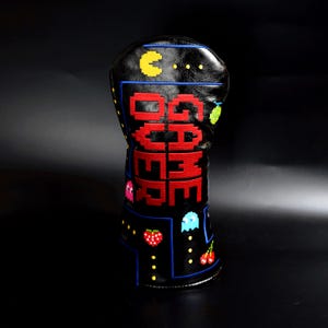 Könnte beinhalten: Schwarze Golfschlägerhaube mit einem pixeligen Videospiel-Design. Die Hülle zeigt das Wort "GAME" in Rot sowie pixelige Charaktere und Symbole. Blaue Nähte umranden das Design.
