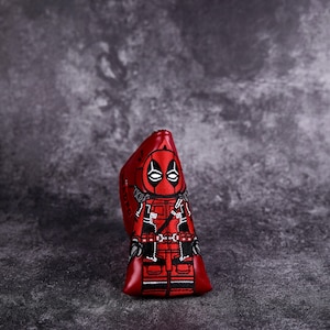 May include: Red golf club head cover featuring a cartoon image of Deadpool in a red and black suit with a mask. The cover has a black and white outline.