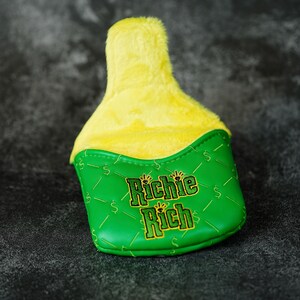 May include: A golf club head cover with a green base and a yellow plush top. The green section features the text "Richie Rich" in gold lettering and dollar signs. The cover is designed to protect golf clubs.