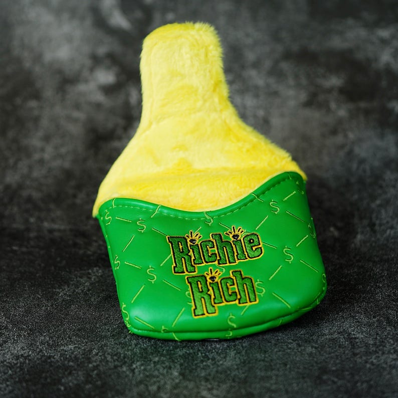 May include: A golf club head cover with a green base and a yellow plush top. The green section features the text "Richie Rich" in gold lettering and dollar signs. The cover is designed to protect golf clubs.