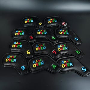 Puede incluir: Un conjunto de fundas para palos de golf negras con la inscripción "GAME OVER" bordada en colores vivos. Cada funda está numerada o con letras, algunas con un diseño de hongo de dibujos animados. Dispuestas sobre una superficie oscura.