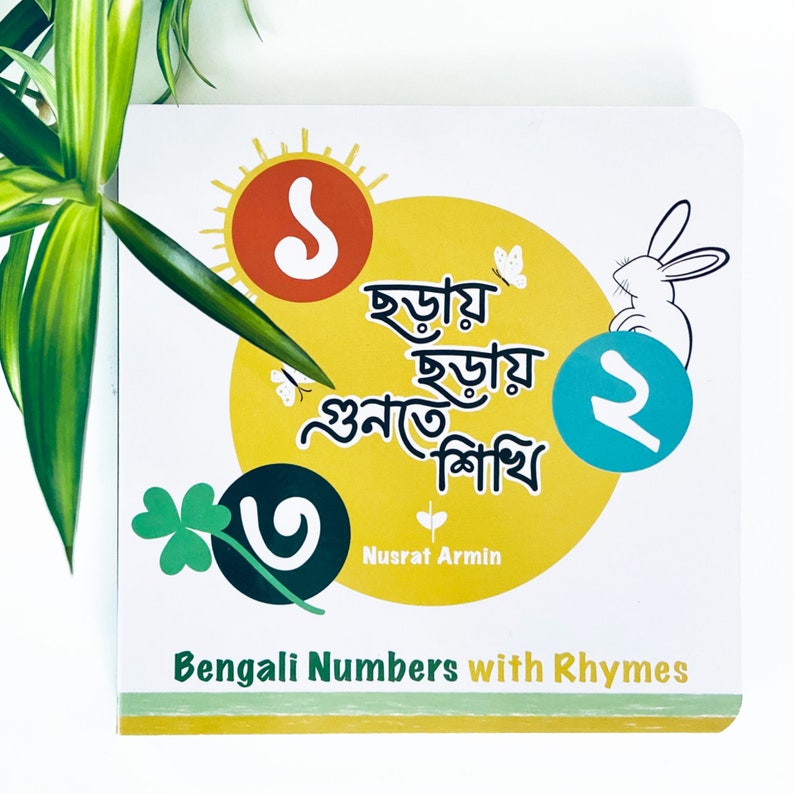 Puede incluir: Un libro colorido con un fondo amarillo y una ilustraci&oacute;n de un conejo blanco. El t&iacute;tulo del libro es "Bengali Numbers with Rhymes" y el texto de la portada est&aacute; en bengal&iacute;. El libro presenta un sol con el n&uacute;mero uno dentro, un tr&eacute;bol con el n&uacute;mero uno dentro y un c&iacute;rculo con el n&uacute;mero dos dentro.