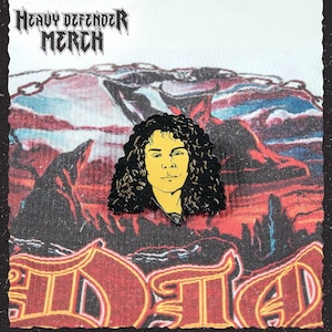 May include: A black and yellow enamel pin depicting a person with long, curly hair. The pin is set against a backdrop of a mountain landscape in red, blue, and white. The words "Heavy Defender Merch" are at the top.
