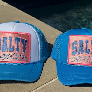 May include: Two blue and white trucker hats with a pink and gold patch that says "Salty" on each hat.