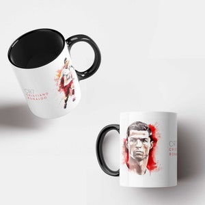 May include: Two white ceramic mugs with black handles. One mug features a watercolour illustration of Cristiano Ronaldo with the text "CR7 CRISTIANO RONALDO". The other mug features a close-up of Cristiano Ronaldo's face with the text "CR7 CRISTIANO RONALDO".