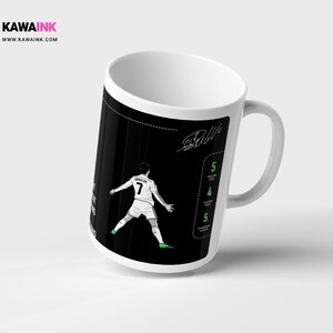 May include: White ceramic mug featuring a black and white illustration of a football player in action. The player is wearing a white jersey with the number 7 on it. The mug has a black background with white text that says "Ronaldo" and "Ball".