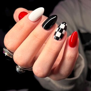 May include: A hand displaying a set of manicured nails. The nails feature a variety of designs, including solid red, white, black, and a black and white checkered pattern. The nails are oval-shaped and have a glossy finish. The hand is adorned with rings.