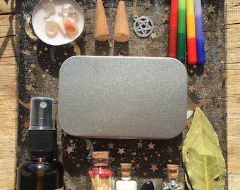 May include: A collection of spiritual items arranged on a black and gold celestial-themed bag. Includes candles, incense cones, crystals, a pentagram charm, small bottles with contents, a spray bottle, a metal tin, a bay leaf, and matches.