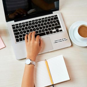 K&ouml;nnte beinhalten: Eine Person tippt auf einem Laptop mit einem Aufkleber auf dem Trackpad, auf dem steht: "Schau mich nicht an, ich habe nicht f&uuml;r ihn gestimmt."