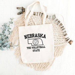 May include: A white canvas tote bag with black text and a volleyball graphic. The text reads "Nebraska The Volleyball State".