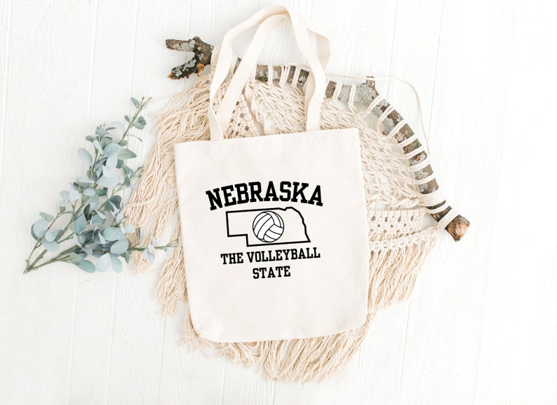 May include: A white canvas tote bag with black text and a volleyball graphic. The text reads "Nebraska The Volleyball State".