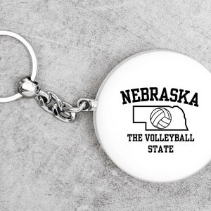 May include: White keychain with a silver chain and a round tag. The tag has a black outline of the state of Nebraska with a volleyball inside and the text "Nebraska The Volleyball State".