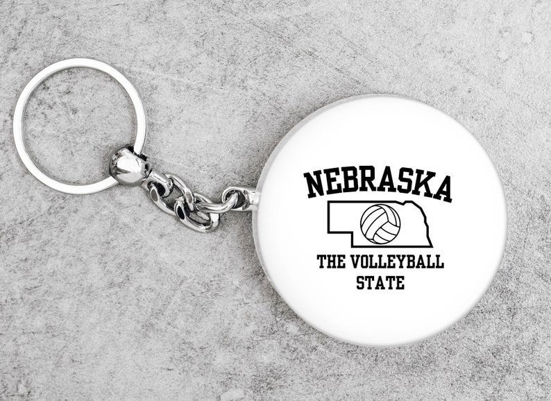 May include: White keychain with a silver chain and a round tag. The tag has a black outline of the state of Nebraska with a volleyball inside and the text "Nebraska The Volleyball State".