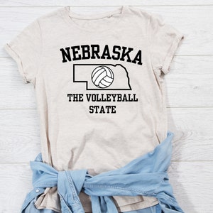 May include: A beige t-shirt with a black outline of the state of Nebraska and a volleyball inside. The text "Nebraska The Volleyball State" is printed on the shirt.