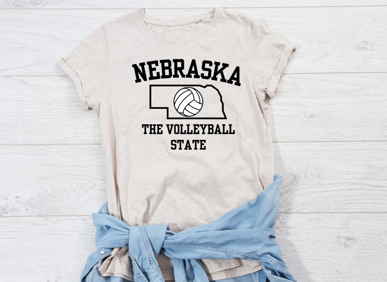 May include: A beige t-shirt with a black outline of the state of Nebraska and a volleyball inside. The text "Nebraska The Volleyball State" is printed on the shirt.